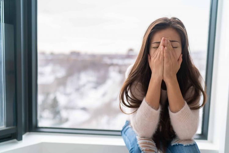 a person holds their face anxiously while sitting in a windowsill