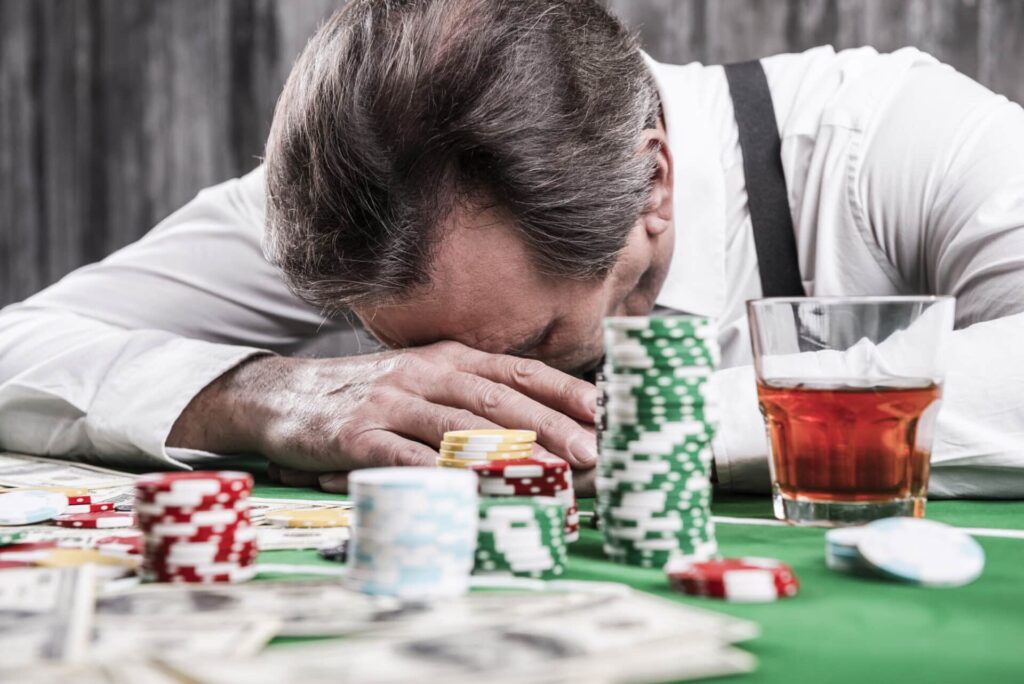 a person puts their head on a poker table behind chips and a glass of liquor