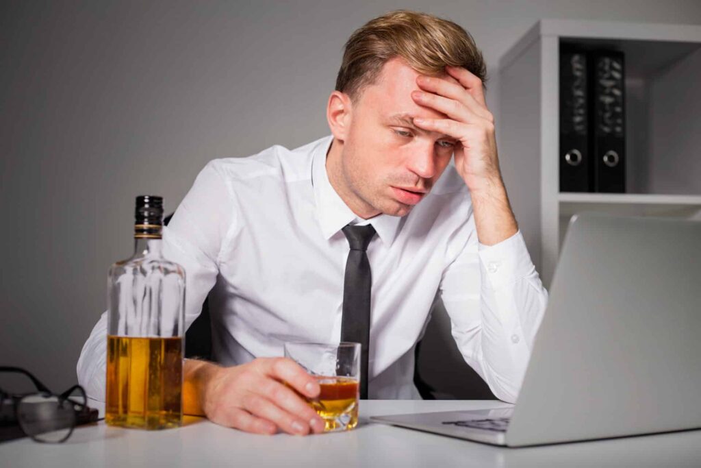 a person looks drunk holding a glass near a liquor bottle as they use a computer