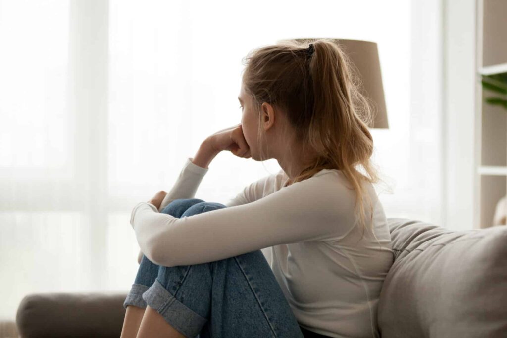 a person sadly clutches their knees on a couch while looking out a window