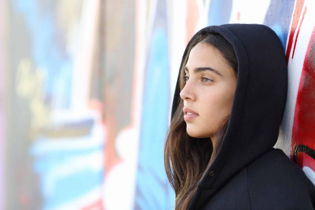 a young person stands with a hoodie on in front of a graffiti wall
