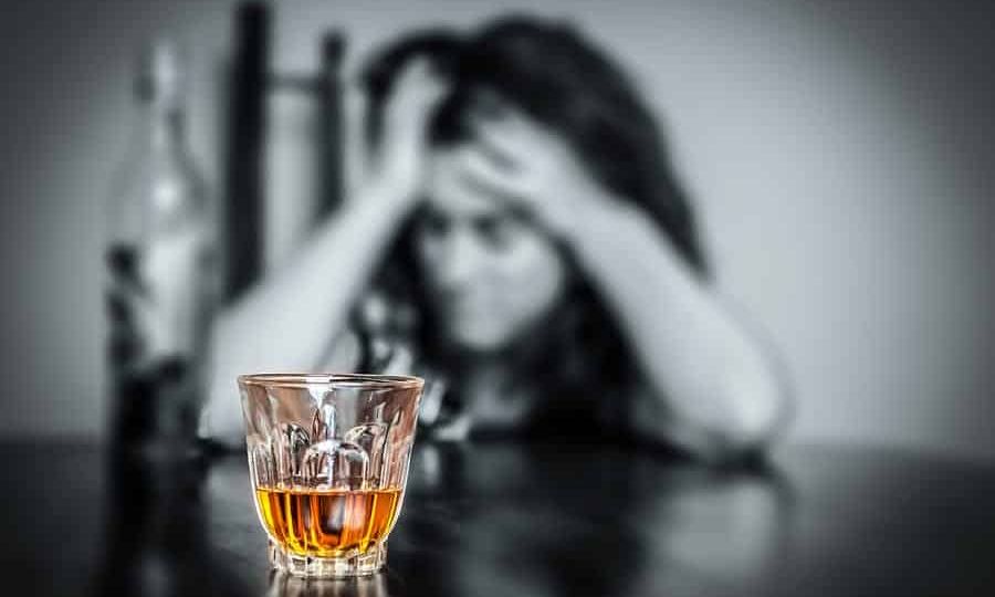 a person holds their head in agony behind a glass of liquor