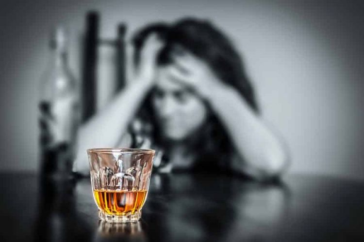 a person holds their head in agony behind a glass of liquor