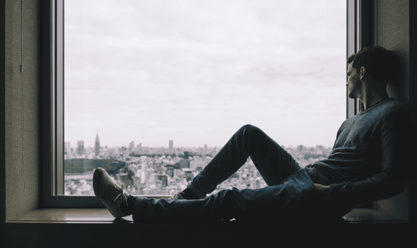 a person sadly looks out a window while sitting in a sill