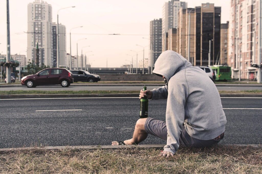 a person holding a beer bottle sits on a curb near a road