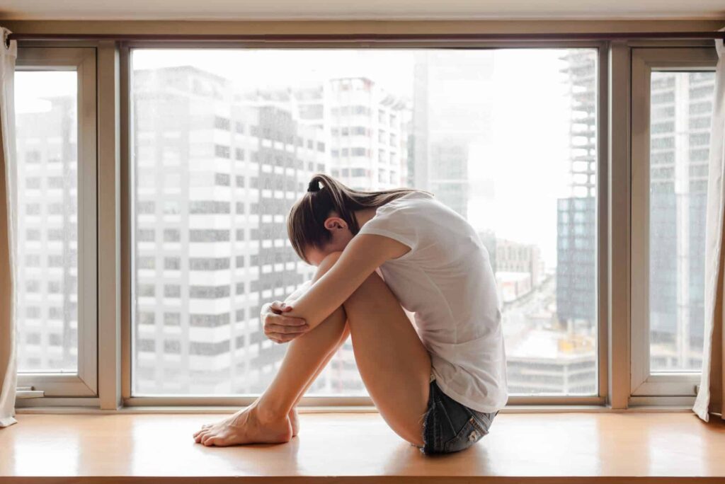 a person sadly holds their head to their knees in a windowsill