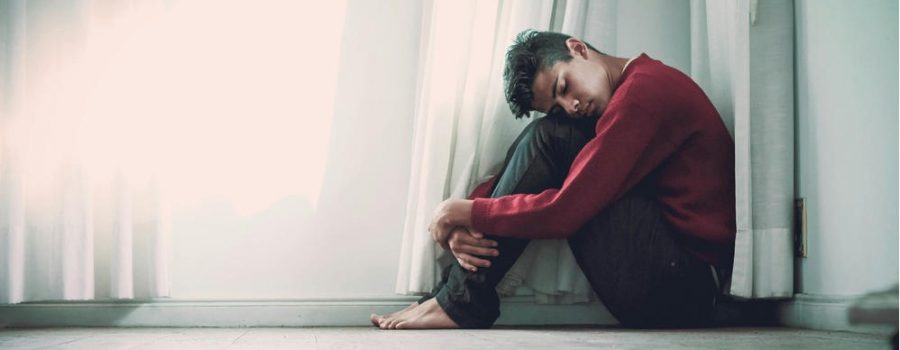 young man sits on the floors with his knees to his chest as he struggles with mental health issues